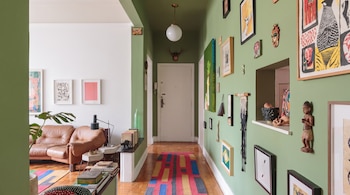 Un pasillo con paredes verdes, cuadros variados y una alfombra multicolor junto a una sala con sofá de cuero marrón y piso de madera.