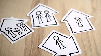 Cuatro recortes de papel blancos en forma de casas, cada uno con dibujos de figuras humanas simples, sobre una tabla de madera clara.