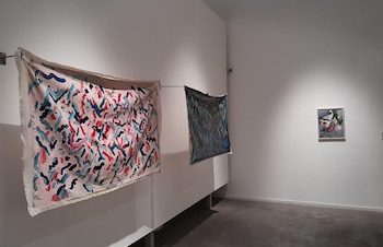 Sala de museo con paredes blancas. Dos obras textiles abstractas cuelgan de una barra. Un cuadro abstracto enmarcado en la pared derecha. El suelo es gris oscuro