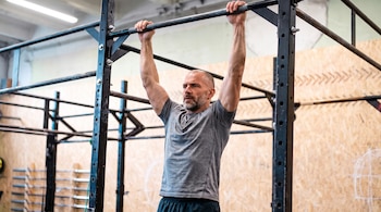 Un hombre de mediana edad, con barba y camiseta gris, se cuelga de una barra de metal horizontal con ambas manos en un gimnasio.