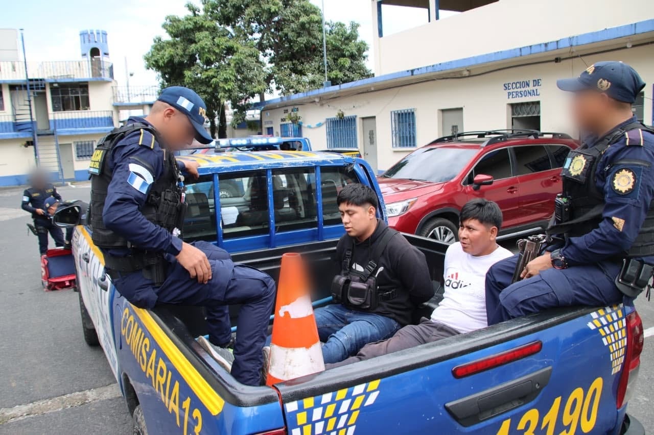 Dos agentes de policía custodian a dos personas detenidas frente a un vehículo oficial junto a armas incautadas y una mochila térmica de reparto en el suelo. (PNCdeGuatemala)