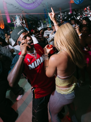 Hombre de tez oscura con gorra, jersey rojo y joyas, hablando por teléfono, junto a mujer rubia con brazo levantado y vaso rojo en un local con gente