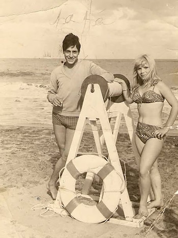 Fotografía antigua de un hombre y una mujer de pie junto a una estructura de madera con un salvavidas en la orilla del mar