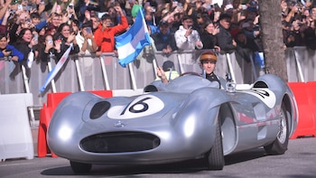 Un piloto joven sonríe dentro de un coche de carreras plateado, con casco de cuero, ondeando una bandera argentina. Detrás, una multitud observa