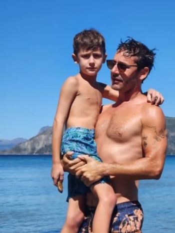 Benjamín Vicuña con gafas de sol sostiene a su hijo Benicio en brazos frente al agua con montañas de fondo