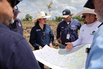 Cinco personas en un campo de tierra cultivada, con árboles pequeños al fondo, miran un mapa grande. Una mujer lo sujeta y un hombre lo señala