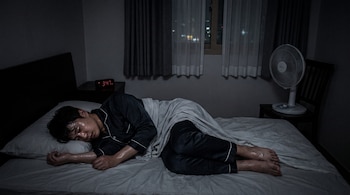 Un hombre con pijama sudado duerme en una cama dentro de una habitación oscura con un reloj digital que marca las 3:47 y un ventilador.