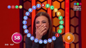 Una mujer sonríe con la mano en la boca frente a una gráfica circular con letras del abecedario en un plató de televisión