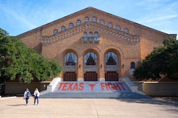 Vista frontal del Gregory Gymnasium de ladrillo con grandes arcos; sus escaleras muestran 'TEXAS FIGHT' y un Longhorn naranja, con dos mujeres caminando