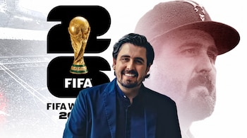 Hombre barbudo sonriente en chaqueta azul, trofeo Copa Mundial y logo FIFA World Cup 26 visibles, con fondo de estadio y una figura difuminada a la derecha.