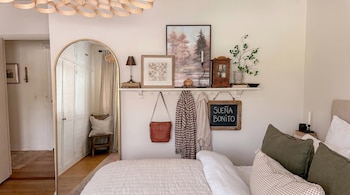 Una cama con cojines verdes en primer plano junto a un espejo arqueado de cuerpo entero y una repisa con cuadros en la pared.