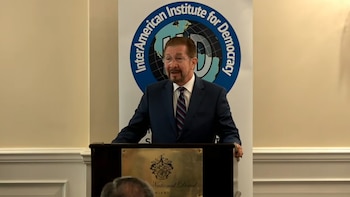 Hombre con traje y gafas detrás de un podio de madera, con una placa de "National Doral Miami". Detrás, un banner con el logo del InterAmerican Institute for Democracy