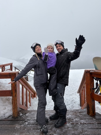 Un hombre y una mujer con traje de esquí posan sobre una plataforma de madera en la nieve sosteniendo a una niña en brazos