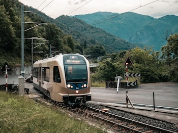 Un tren blanco y amarillo cruza un puente de acero sobre un río en un valle montañoso cubierto de densa vegetación verde, con nubes bajas en las cimas