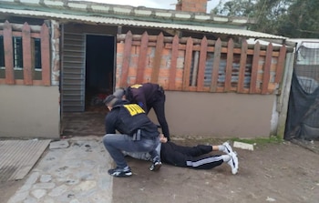 Dos agentes con chalecos de la DDI sujetan a un hombre tendido en el suelo frente a una casa con fachada rústica y un cerco de madera