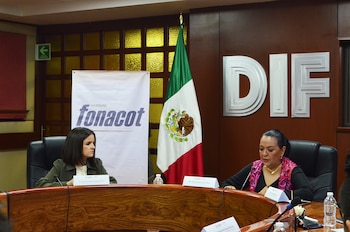 Dos mujeres sentadas frente a una mesa de madera. Detrás hay un cartel de Fonacot, la bandera de México y el logo "DIF" en una pared marrón
