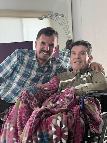 Dos hombres sonrientes, uno con camisa a cuadros y otro en silla de ruedas con polo marrón y manta de flores, posan en un interior