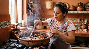 Una mujer viste blusa bordada y revuelve chiles secos con una cuchara de madera dentro de una cazuela de barro sobre la estufa.