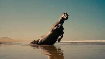 Vista lateral de una gran escultura metálica con forma de caballo de guerra, parcialmente sumergida en el agua y la arena de una playa