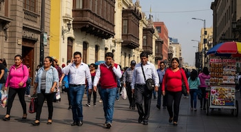 Personas caminan por una calle concurrida en Lima, flanqueada por edificios históricos con balcones de madera, y un puesto de comida ambulante.