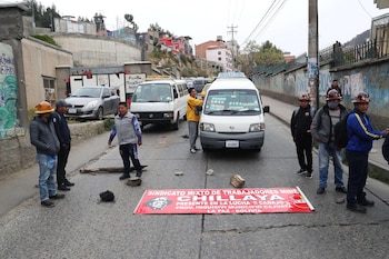 Una calle está bloqueada por hombres, rocas, madera y un cartel rojo. Se ven dos furgonetas blancas y un coche gris detenidos. Edificios y colinas en el fondo