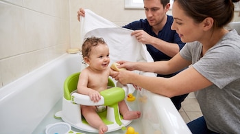 Un bebé sentado en un asiento verde de plástico dentro de una bañera con agua junto a dos adultos, una esponja y patos de goma.