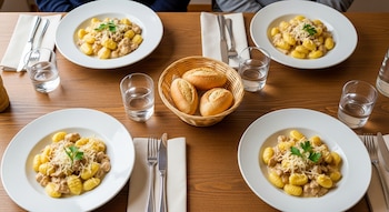 Cuatro platos de ñoquis con salsa de pollo a la crema, queso rallado y perejil en una mesa de madera con cubiertos, servilletas, vasos y una panera con pan.