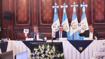 Delegados sentados en una mesa en una sala formal, levantan carteles blancos con la palabra 'SÍ'. Un fotógrafo captura la escena al fondo