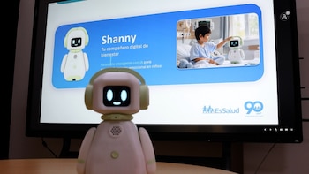 Un robot rosado con auriculares verdes en primer plano frente a una pantalla que muestra información del robot Shany y la foto de un niño interactuando con otro robot en un hospital
