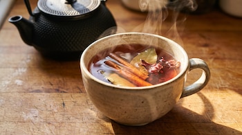 Taza de bebida caliente rojiza con vapor, ramitas de canela, hojas de laurel y flores secas de jamaica, junto a una tetera negra sobre un mostrador de madera.