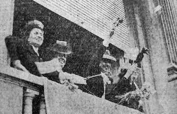 Fotografía en blanco y negro de varias personas asomadas a un balcón agitando sombreros y saludando