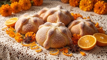 Panes espolvoreados con azúcar sobre un mantel blanco tejido junto a flores anaranjadas de cempasúchil, rodajas de naranja y anís estrella.