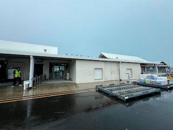 Edificio del Aeropuerto Internacional Bonriki con letrero azul, un hombre con chaleco amarillo, pavimento mojado, dos carros de carga, cielo gris