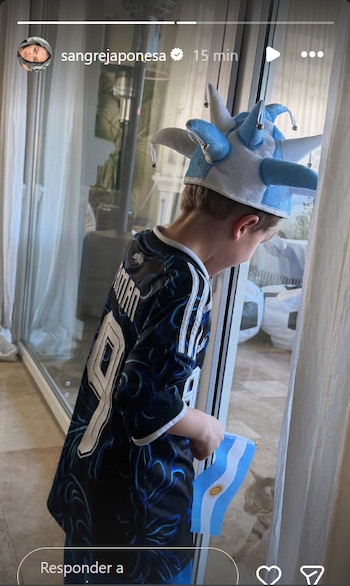Niño de cabello rubio con camiseta azul y blanca, gorro de bufón celeste y blanco, y bandera argentina. Mira por una ventana junto a cortinas blancas