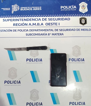 Un cuchillo de sierra con mango negro y un teléfono celular oscuro sobre una superficie blanca con insignias de la Policía de la Provincia de Buenos Aires