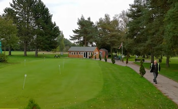 Campo de práctica de golf verde con banderas blancas, un edificio de madera, un hombre con chaleco, un sendero y una bandera británica