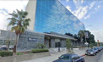 Un edificio de cristal azul de varios pisos con la señal del Departamento de Policía de Long Beach, flanqueado por palmeras y una calle con coches estacionados