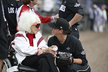 Una mujer mayor con cabello blanco y gafas de sol rojas en silla de ruedas observa a una jugadora de béisbol agachada que sostiene una pelota y un guante