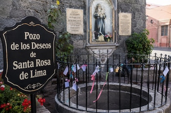 Un pozo de piedra rodeado por una reja con papeles atados, un cartel con texto y una hornacina con una escultura religiosa al fondo