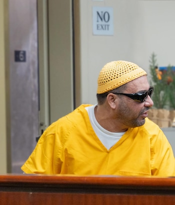 Un hombre con gorro de ganchillo amarillo y gafas de sol, vestido con una camisa amarilla de manga corta y una camiseta blanca, en un interior