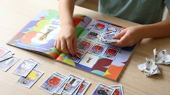 Manos de un niño pegando una lámina de fútbol en un álbum Panini abierto para el Mundial 2026 sobre una mesa de madera clara, con más láminas alrededor.