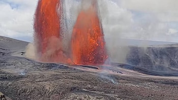 Múltiples fuentes de lava de color naranja brillante y rojo se elevan desde el suelo volcánico oscuro, con humo gris y un cielo nublado de fondo