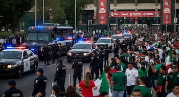 Varios coches y un vehículo blindado de policía, con luces encendidas, junto a agentes. Una multitud lleva camisetas verdes y banderas de México.