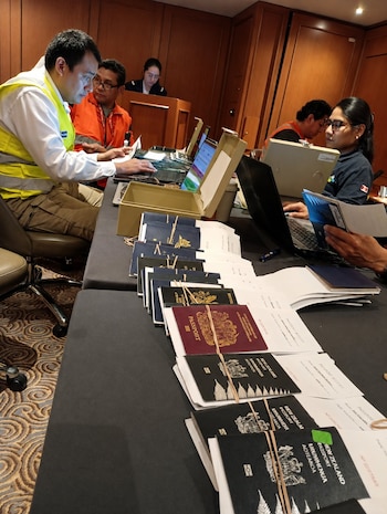 Varias personas sentadas frente a computadoras portátiles, con pilas de pasaportes atados y documentos sobre una mesa en una sala