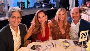 Cuatro invitados sonrientes, dos hombres y dos mujeres, sentados en una mesa de gala con comida y copas de vino. Un escenario con equipo musical al fondo