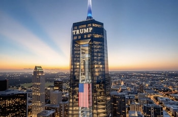 Un rascacielos moderno con el nombre 'TRUMP' iluminado y una gran bandera de Estados Unidos en su fachada, con un horizonte urbano al atardecer
