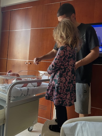 Un hombre y una niña de cabello rubio miran a un bebé recostado en una cuna transparente de hospital, con fondo de pared de madera
