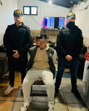 Un hombre con una sudadera color crema y chaqueta oscura está sentado y esposado, flanqueado por dos agentes de policía con uniformes negros y chalecos antibalas