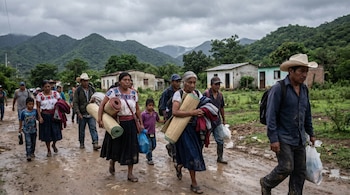 Un grupo de personas indígenas, incluyendo mujeres y niños, camina por un camino de tierra embarrado. Llevan esteras enrolladas y bolsas. Al fondo, se ven casas y montañas verdes bajo un cielo nublado.