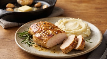 Pechuga de pollo rebanada sobre un plato cerámico con puré de papas, romero y una sartén de hierro con mantequilla al fondo.